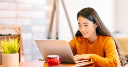 一个穿着橙色毛衣的女人坐在桌子旁，专注地使用笔记本电脑. 